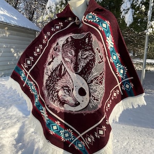 May include: A burgundy poncho with a wolf design in the center, featuring a yin-yang symbol. The poncho has a fringed hem and geometric patterns in teal, white, and red. A button closure is visible at the collar.