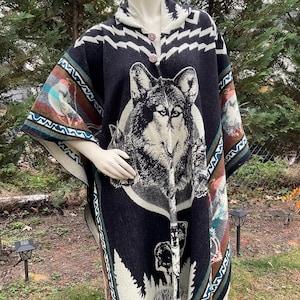 Op de afbeelding: Een zwarte poncho met capuchon en een wolfontwerp. De poncho heeft een grote wolfskop in het midden, omgeven door een witte cirkel. De randen zijn afgewerkt met kleurrijke patronen en franjes.