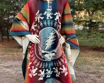 May include: A colorful poncho with a hood and fringe, featuring a design with dancing bears and a skull with a lightning bolt. The poncho has a mix of red, orange, blue, and green stripes and patterns.