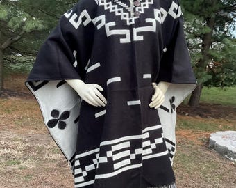 May include: Black and white poncho with a hood and geometric patterns. The poncho has a fringed hem and a button closure at the neck. The design includes white shapes on a black background, with a contrasting gray and white interior.