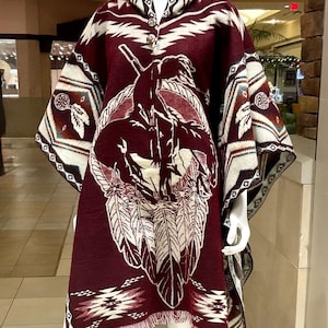 May include: A burgundy poncho with a Native American design, displayed on a mannequin. The poncho features a central graphic of a Native American figure on horseback, surrounded by feathers. The trim and hood have geometric patterns in white, brown, and burgundy.