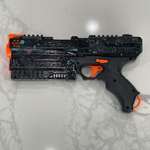 Può includere: Pistola giocattolo nera e arancione con un design a splatter paint. La pistola ha un'impugnatura nera e una canna nera con un grilletto arancione. La pistola ha un caricatore nero e arancione.