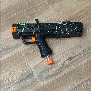 May include: A black and green toy gun with an orange barrel and a black handle. The gun has a splatter paint design.