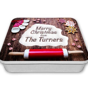 Op de afbeelding: Een witte metalen koekjestrommel met een houtnerfdesign. De trommel heeft een rode deegroller, een kom met wit meel met de woorden "Merry Christmas from The Turners" erop geschreven, en verschillende kerstthema-koekjesvormen en snoepjes.