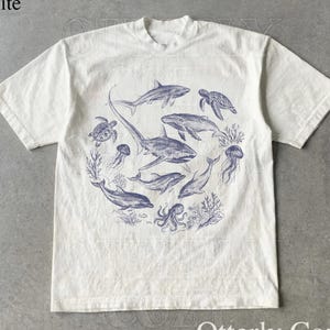 May include: White t-shirt featuring a blue illustration of various sea creatures, including sharks, dolphins, and turtles, arranged in a circular pattern. The text "Otterly Cozy" is printed at the bottom of the shirt.