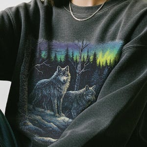 May include: Dark gray sweatshirt featuring a graphic of two wolves in a forest setting with an aurora borealis effect. The text "Otterly Cozy" is printed at the bottom. The sweatshirt is worn by a person.
