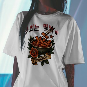 Puede incluir: Camiseta blanca con un diseño de tteokbokki, un popular plato callejero coreano. El diseño incluye un cuenco con el plato, palillos y el texto "TTEOKBOKKI" en una pancarta. La camiseta tiene cuello redondo clásico.
