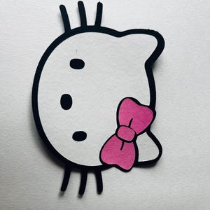 May include: Black and white outline of a cartoon cat with a pink bow. The cat has three whiskers on each side and three black dots for eyes.
