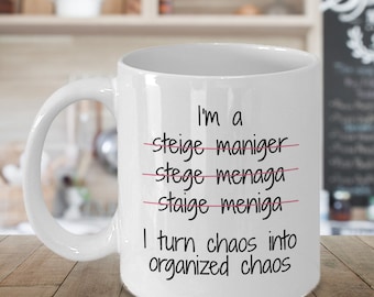 May include: White ceramic coffee mug with black text that reads "I'm a stage manager I turn chaos into organized chaos". The words "stage manager" are crossed out with a pink line.