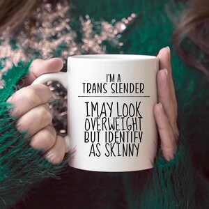 May include: White ceramic mug with black text that reads "I'm a Trans Slender I may look overweight but identify as skinny".