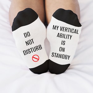 May include: A pair of white socks with black trim. One sock says "Do Not Disturb" with a red circle and slash. The other sock says "My Vertical Ability Is On Standby".