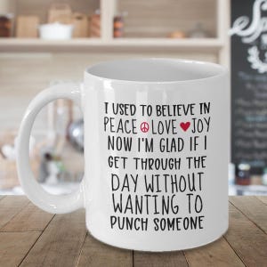 May include: White ceramic mug with a large handle. The mug features black text that reads: "I USED TO BELIEVE IN PEACE LOVE JOY NOW I'M GLAD IF I GET THROUGH THE DAY WITHOUT WANTING TO PUNCH SOMEONE."