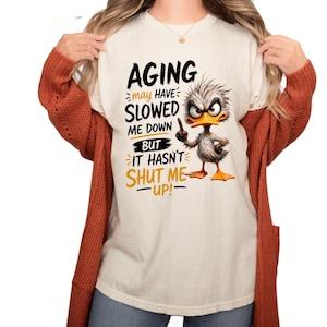 Op de afbeelding: Een crèmekleurig t-shirt met een afbeelding van een chagrijnige, warrige eend met oranje voeten en snavel. De tekst luidt: AGING may have SLOWED ME DOWN BUT IT HASN'T SHUT ME UP! De eend wijst met zijn vleugel uitdagend omhoog.