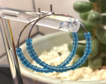May include: A pair of hoop earrings made with light blue beads. The earrings are displayed on a clear plastic stand.