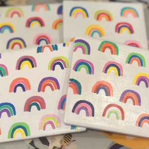 Puede incluir: Cuatro posavasos cuadrados blancos con diseños de arcoíris pintados a mano. Cada posavasos presenta un patrón repetitivo de arcoíris coloridos en tonos de rojo, naranja, amarillo, verde, azul y morado. Los posavasos están dispuestos de forma ligeramente superpuesta.