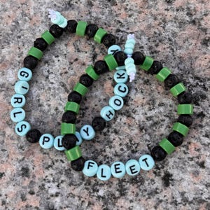 Peut inclure: Deux bracelets en perles avec des perles de lave noires et des perles de lettres vertes et bleu clair. Les bracelets épellent "Girls Play Hockey" et "Fleet".