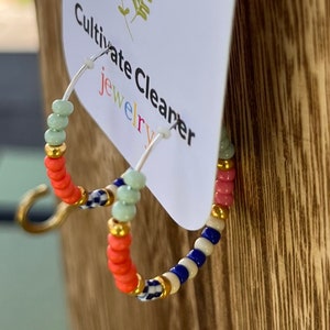 May include: A pair of hoop earrings with colorful beads. The beads are in a pattern of pink, blue, white, and green. The earrings are on a white card with the text "Cultivate Cleaner Jewelry".