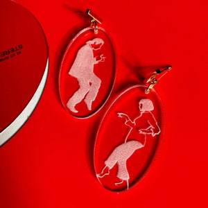 May include: A pair of oval-shaped earrings with a white silhouette of a person on each earring. The earrings are made of clear acrylic and have gold-colored hooks.