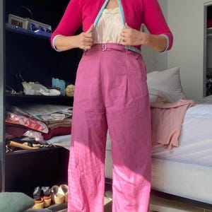 May include: A woman's outfit featuring a fuchsia pink cardigan with light blue trim, a cream-colored top, and high-waisted, wide-leg fuchsia pink pants. The outfit is accessorized with a belt. The background includes a closet and a bed.