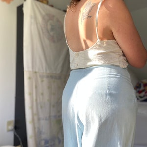 May include: A person wearing a white tank top and light blue wide-leg pants. The person has a tattoo on their back.