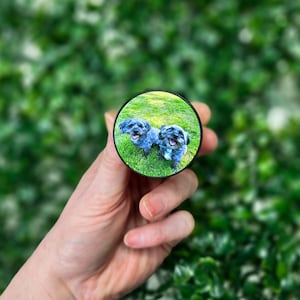 May include: A black phone pop socket with a photo of two black dogs on a green grassy field.