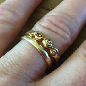 Pode incluir: Um close-up de uma mão usando dois anéis de ouro. O anel superior tem um design curvo com pequenas pedras vermelhas e transparentes. O segundo anel é uma simples banda de ouro. O fundo é uma superfície de madeira.