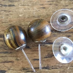 Könnte beinhalten: Ein Paar Ohrstecker mit kugelförmigen Tigerauge-Edelsteinen in braun-gold gestreiften Mustern. Die Ohrringe haben silberfarbene Metallstifte und zwei durchsichtige, kreisförmige Kunststoff-Ohrsteckerverschlüsse für einen sicheren Halt.
