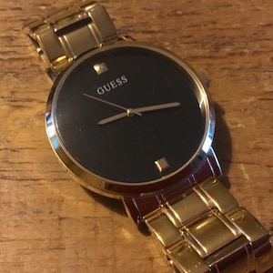 May include: A gold-coloured Guess wristwatch with a black watch face. The watch features gold hands and hour markers, and the brand name "GUESS" is printed in white. The watch strap is also gold-coloured.