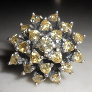 Op de afbeelding: Zilveren ring met gele edelstenen in een bloemmotief. Een ronde steen in het midden, omringd door hartvormige edelstenen. De zilveren zetting contrasteert met de warme, gouden kleur van de stenen.