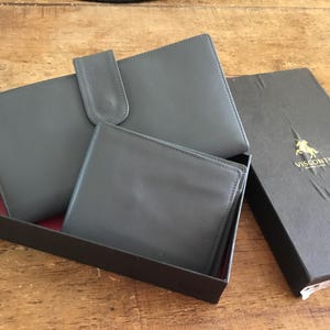 Op de afbeelding: Een grijze lederen portemonneeset bestaande uit een grote rechthoekige portemonnee met drukknoopsluiting en een kleinere opvouwbare portemonnee. De items worden gepresenteerd in een zwarte doos met het gouden Visconti-logo en een paardenembleem.