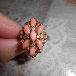 May include: A gold-toned ring featuring a cluster of oval, light pink gemstones surrounded by smaller clear stones. The ring has a detailed, floral-like design around the gemstones, creating a vintage aesthetic. The ring is a piece of jewelry.