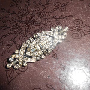 May include: A vintage silver brooch adorned with clear rhinestones. The brooch features an elongated, symmetrical design with a central rectangular stone and smaller, round stones. It rests on a patterned, dark brown surface.