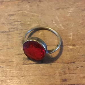 Peut inclure: Une bague dotée d'une pierre rouge circulaire à facettes, montée sur un anneau métallique fin et simple. La pierre est maintenue par un serti clos avec un détail dentelé subtil sur le pourtour, révélant une couleur rouge vive et réfléchissante.