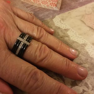 May include: A black ring with a silver band and a row of small, clear stones. The ring has a cross-like design with a band of clear stones. The ring is on a person's finger.