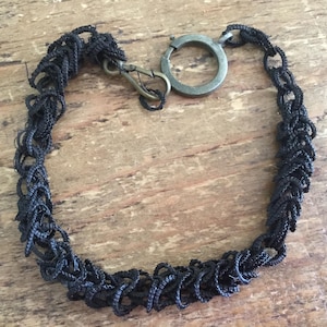May include: A black woven cord bracelet featuring a textured, chain-like pattern. The bracelet is secured with a metallic bronze-toned lobster claw clasp and a circular ring connector, creating a continuous loop design with a dark, matte finish.