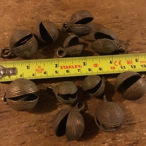 Puede incluir: Una colección de once pequeñas campanas de metal redondas de color bronce con texturas estriadas horizontales y una ranura vertical. Una cinta métrica amarilla Stanley se coloca sobre las campanas, mostrando tamaños de 1,5 a 2,5 cm.