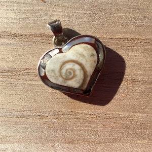 Peut inclure: Pendentif en forme de cœur avec une bélière argentée. Le pendentif présente un motif en spirale au centre, entouré d'une bordure marron foncé. Le fond est une surface en bois brun clair.