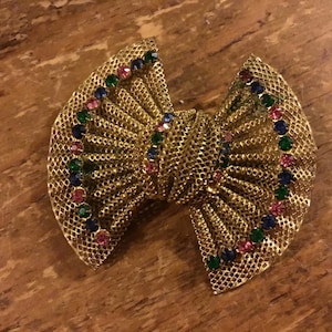 Pode incluir: Um broche de metal dourado em forma de laço com uma superfície de malha texturizada. O laço é decorado com uma fileira curva de pequenos strass redondos alternando entre verde, azul e rosa em cada lado e uma faixa central correspondente.