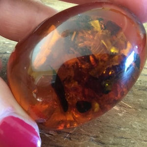 Puede incluir: Una pieza de ámbar translúcido pulido de forma ovalada con un tono naranja miel intenso. La piedra presenta inclusiones internas visibles, incluyendo fragmentos orgánicos oscuros y motas de color marrón dorado suspendidas en la resina brillante.