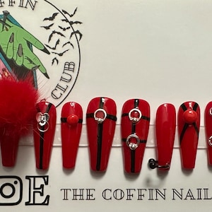 Puede incluir: Conjunto de diez u&ntilde;as postizas rojas en forma de ata&uacute;d con rayas negras y detalles plateados. Las u&ntilde;as presentan una variedad de dise&ntilde;os, incluyendo anillos, tachuelas y un peque&ntilde;o pomp&oacute;n rojo.