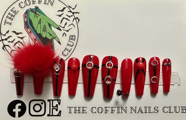 Puede incluir: Conjunto de diez u&ntilde;as postizas rojas en forma de ata&uacute;d con rayas negras y detalles plateados. Las u&ntilde;as presentan una variedad de dise&ntilde;os, incluyendo anillos, tachuelas y un peque&ntilde;o pomp&oacute;n rojo.