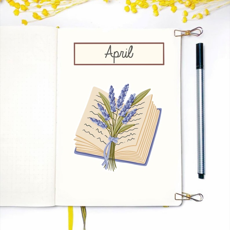 May include: An open notebook with the word "April" in a brown rectangle. A drawing of a book with a lavender bouquet tied with a blue ribbon is on the right page. A silver pen and two gold paper clips are also visible.
