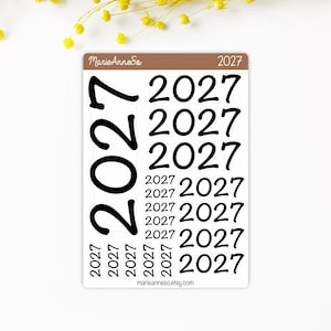 Könnte beinhalten: Ein rechteckiger Aufkleberbogen mit dem Text 2027 in verschiedenen Größen und Ausrichtungen. Die braune Kopfzeile enthält MarieAnneCo und 2027. Der Bogen enthält eine große vertikale 2027, drei mittlere 2027 und mehrere kleinere 2027.