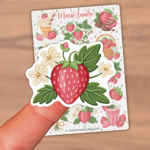May include: A strawberry sticker with a large red strawberry, green leaves, and white flowers. The sticker is held by a finger. The background features a sheet with strawberry-themed illustrations, including ice cream and chocolate.