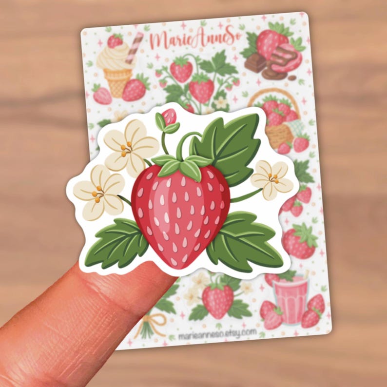 May include: A strawberry sticker with a large red strawberry, green leaves, and white flowers. The sticker is held by a finger. The background features a sheet with strawberry-themed illustrations, including ice cream and chocolate.