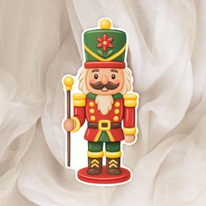 Peut inclure: Autocollant représentant un soldat casse-noisette de dessin animé. Le casse-noisette porte un chapeau vert avec une fleur rouge, une moustache et des cheveux blancs, et un uniforme rouge avec des détails dorés. Il tient un bâton doré et se tient sur une base rouge.