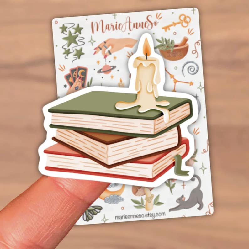 Puede incluir: Una pegatina troquelada que muestra una pila de tres libros en verde, marr&oacute;n y rojo con una vela de color crema derriti&eacute;ndose encima. La pegatina est&aacute; sobre una hoja con varias ilustraciones m&iacute;sticas y el texto MarieAnneSo.