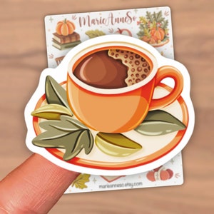 K&ouml;nnte beinhalten: Ein Aufkleber mit einer Cartoon-Illustration einer Tasse Kaffee mit brauner Fl&uuml;ssigkeit und Schaum auf einer Untertasse mit gr&uuml;nen Bl&auml;ttern. Der Aufkleber befindet sich auf einem Finger.