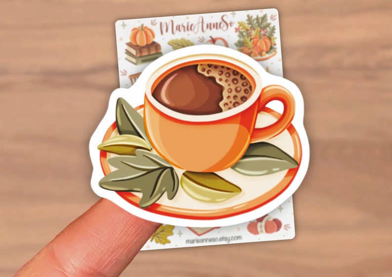 K&ouml;nnte beinhalten: Ein Aufkleber mit einer Cartoon-Illustration einer Tasse Kaffee mit brauner Fl&uuml;ssigkeit und Schaum auf einer Untertasse mit gr&uuml;nen Bl&auml;ttern. Der Aufkleber befindet sich auf einem Finger.