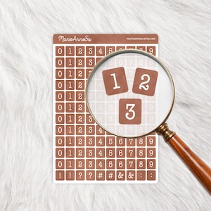 Peut inclure: Une feuille d'autocollants de chiffres bruns avec des chiffres et des symboles blancs. Une loupe se concentre sur les chiffres 1, 2 et 3. Les autocollants incluent les chiffres 0-9 et des symboles. Le nom et le site Web de la boutique Etsy sont visibles.